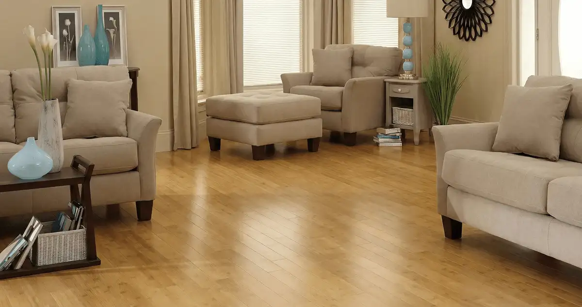 A living room with professionally installed bamboo flooring, beige sofas, a matching ottoman, a side table with a blue lamp, vases, framed pictures, and large windows with blinds and curtains in Aurora, CO